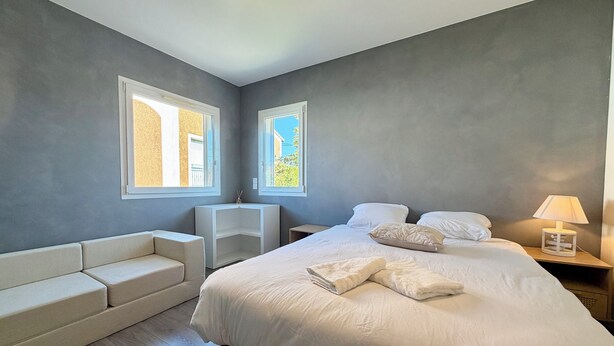 A bedroom with a large bed, a bedside table with a lamp, a small white chair, and a built-in bench along the wall.