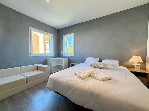 A bedroom with a large bed, a bedside table with a lamp, a small white chair, and a built-in bench along the wall.
