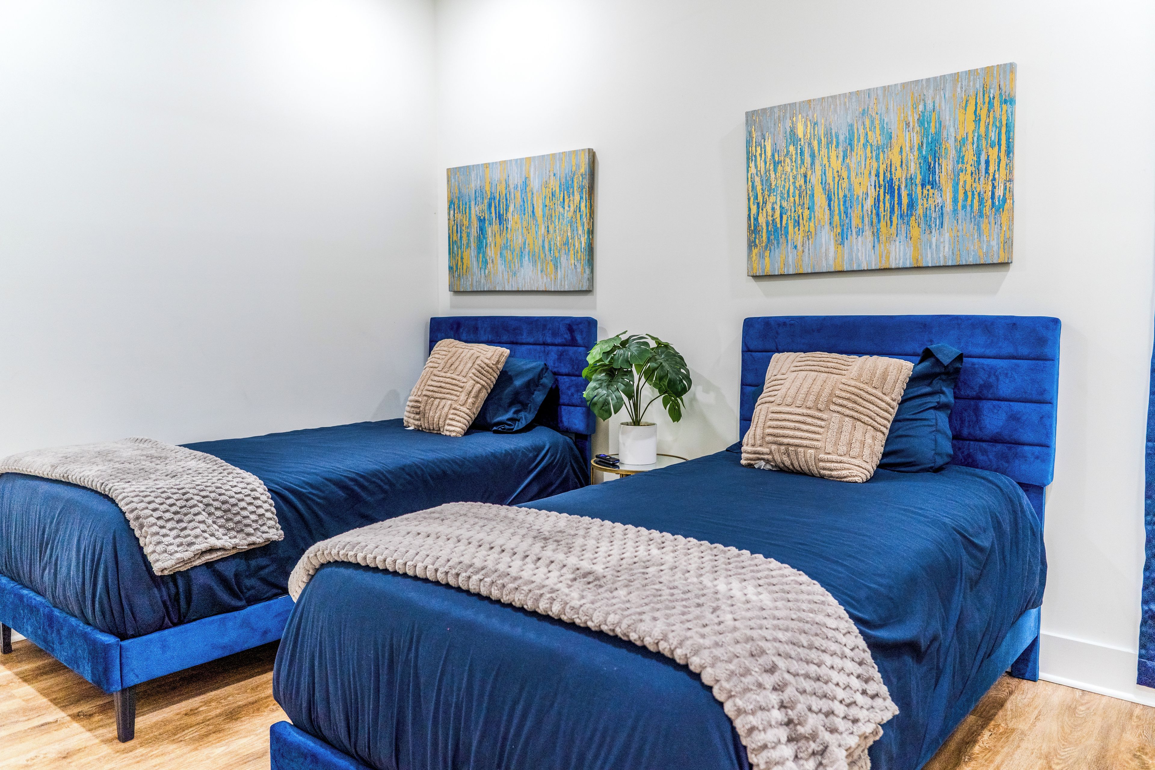 Une chambre avec deux lits, chacun doté d’une tête de lit bleue et d’une couverture beige. Deux tableaux abstraits sont accrochés au mur, au-dessus des lits.