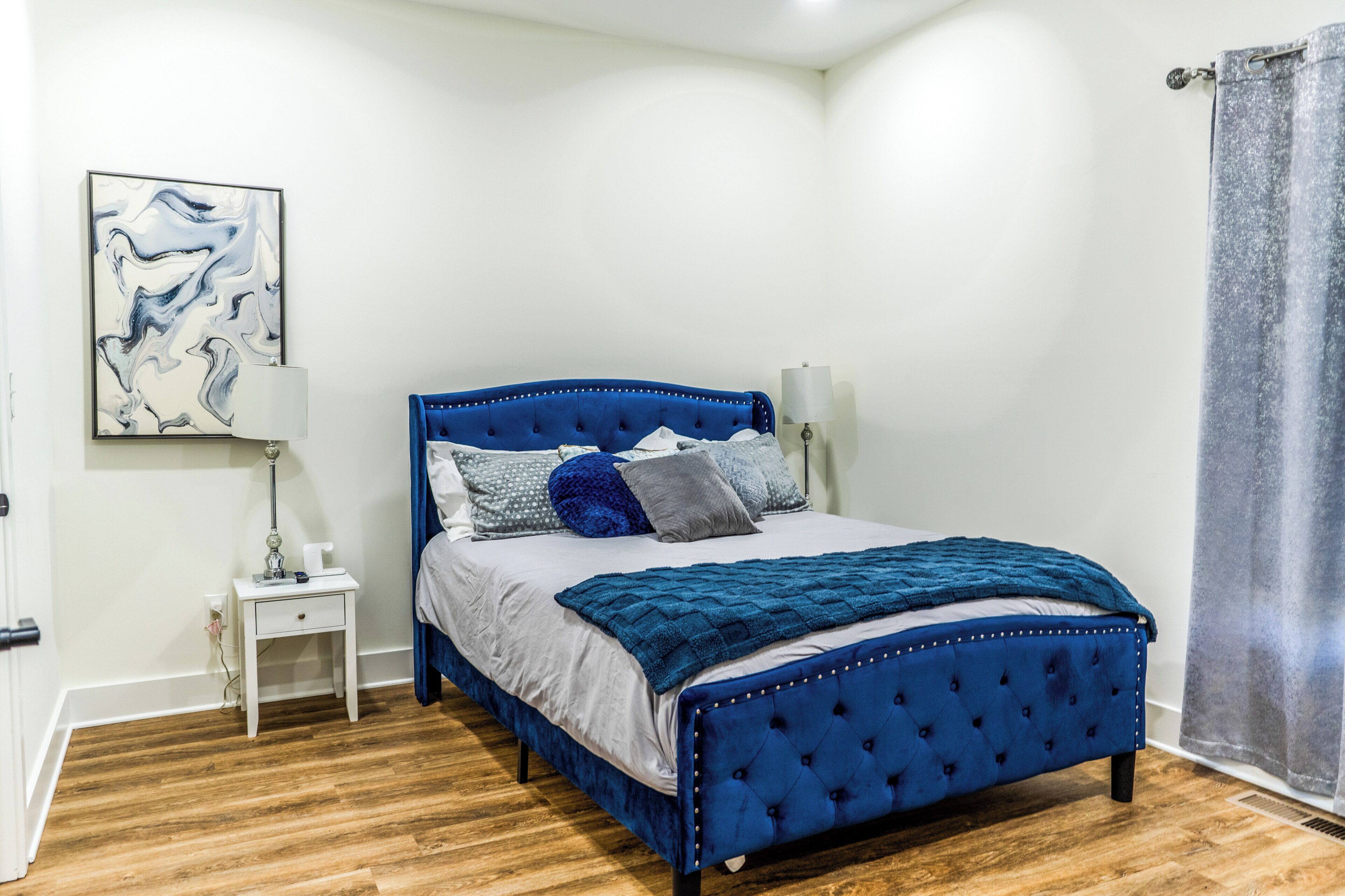 Ein Schlafzimmer mit einem blau gesteppten Bett, einem weißen Nachttisch, einer Lampe und einem Bild an der Wand.