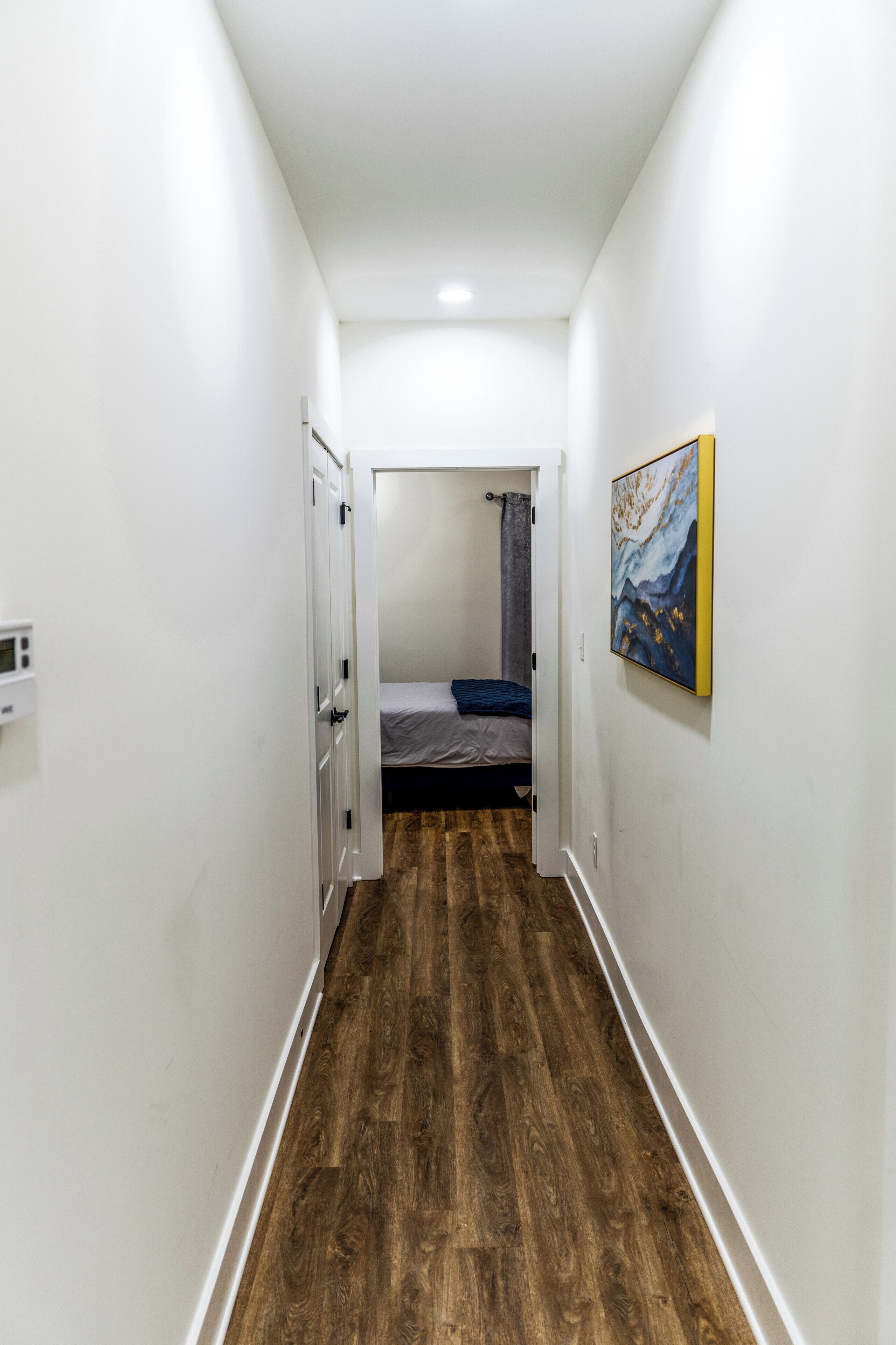 Un estrecho pasillo con suelo de madera que conduce a un dormitorio con cama y un cuadro en la pared.