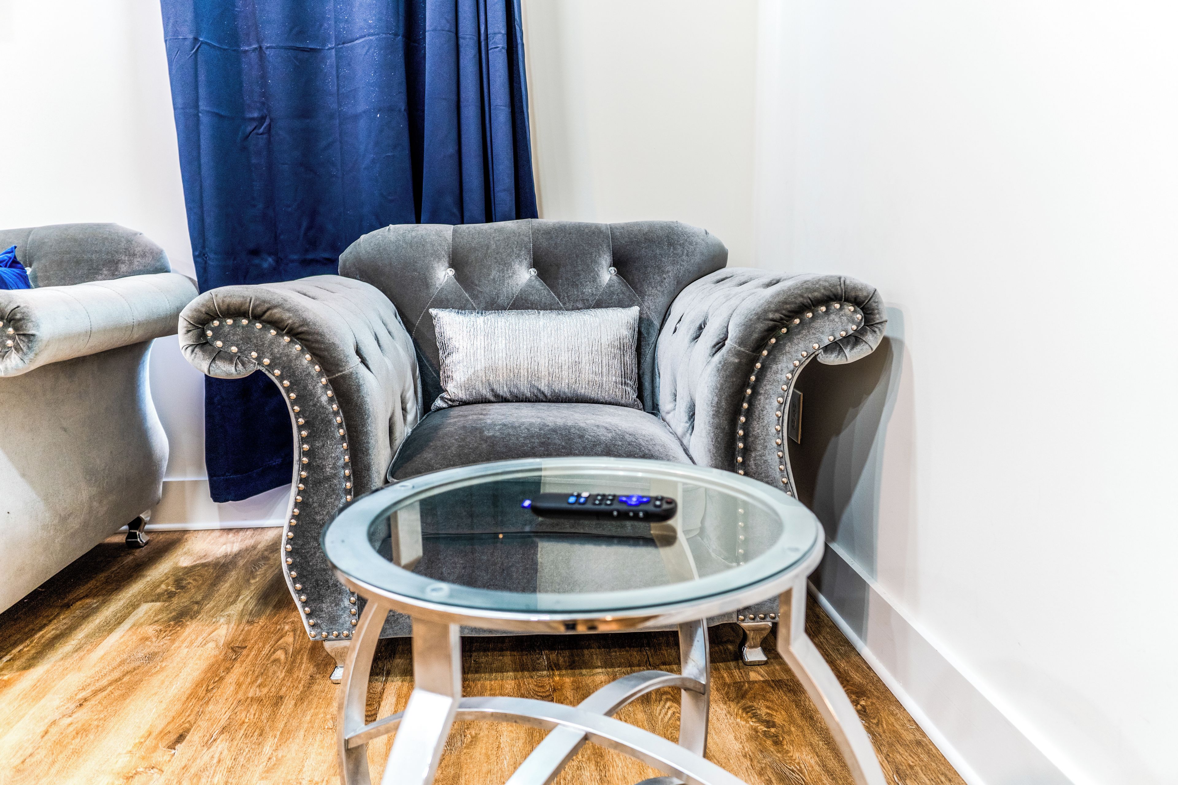 Un sillón gris con respaldo capitonado y botones decorativos, ubicado junto a una mesa de centro de vidrio con un control remoto encima.