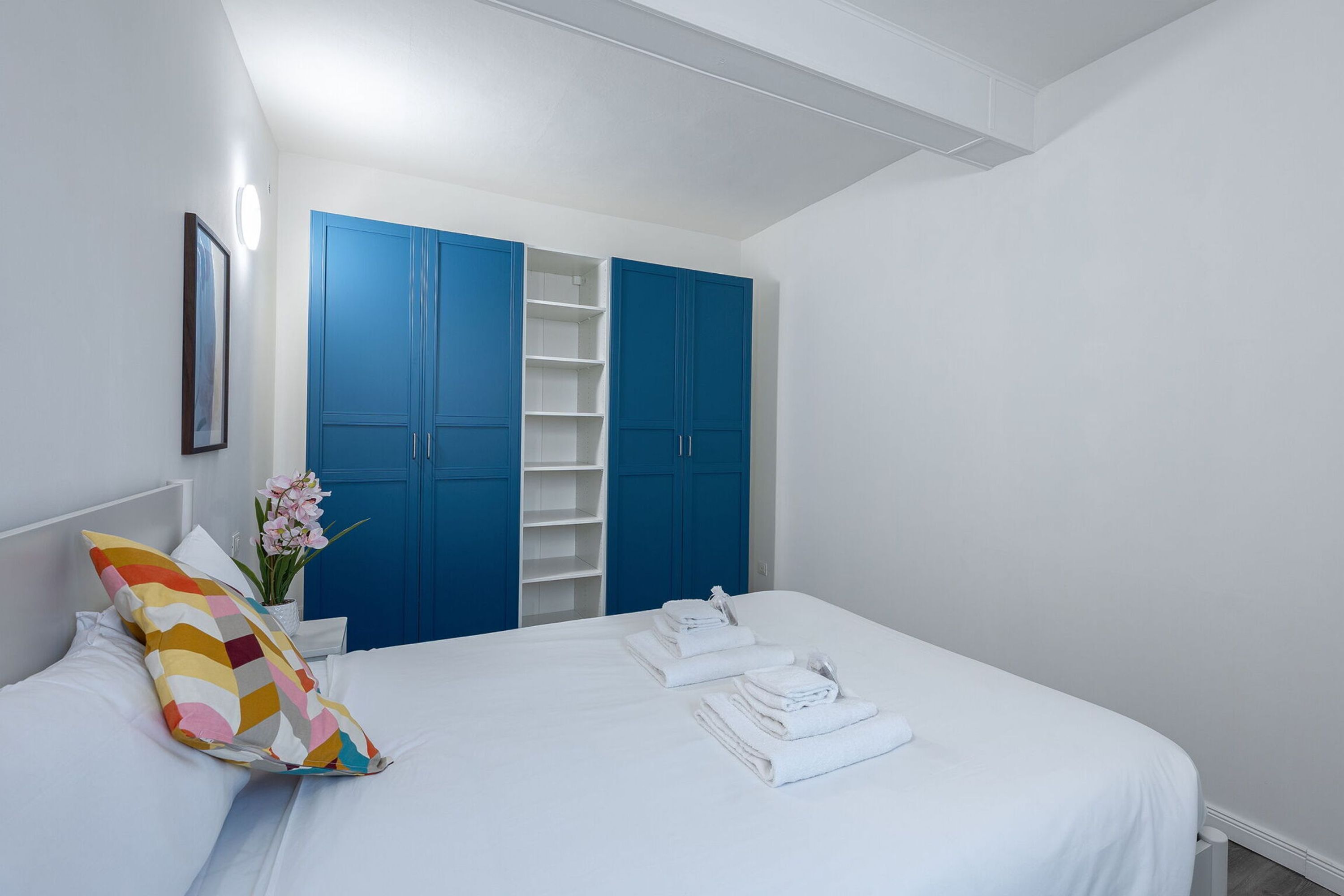 Un dormitorio con cama, un cojín colorido, un jarrón con flores y un armario azul.