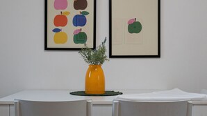 Una mesa de comedor con sillas, una obra de arte enmarco con manzanas coloridas y un jarrón con follaje verde.