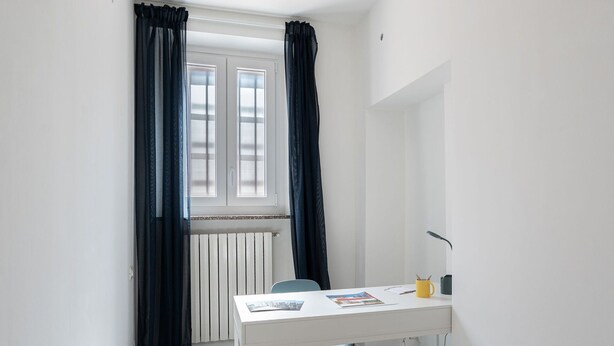 Una habitación pequeña y luminosa con un escritorio blanco, una silla, un radiador y una ventana con cortinas.