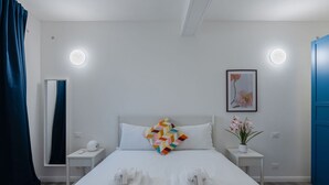 Habitación de hotel con cama, dos mesitas de noche, una lámpara de noche, un reloj de pared, un cuadro enmarco, una planta en maceta y una puerta azul.