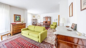 Uma sala de estar com um sofá verde, um armário de madeira, um espaço de refeições com mesa e cadeiras, e uma lareira de mármore.