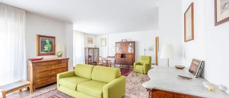 A living room with a green sofa, a wooden cabinet, a dining area with a table and chairs, and a marble fireplace.