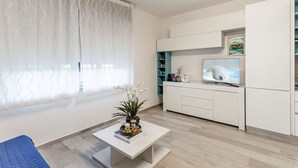 Una sala moderna con un sofá blanco, una mesita de centro con libros y una planta, una cocina con armarios blancos, un televisor y un ventanal con cortinas translúcidas.