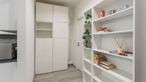 Un pasillo con un armario blanco, una estantería blanca con libros y objetos decorativos, y una puerta que da a otra habitación.