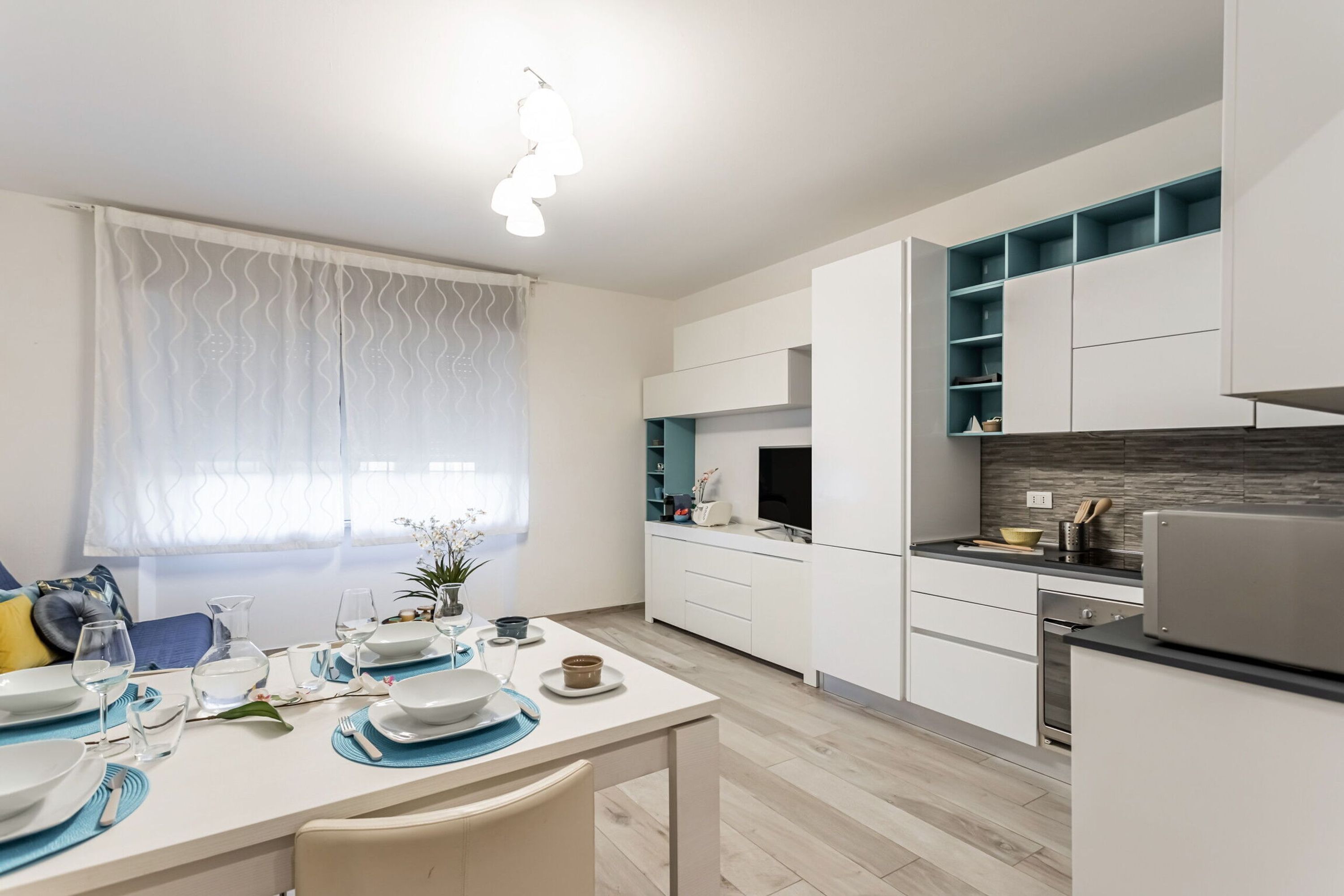 Una cocina moderna con armarios blancos, un comedor con mesa para dos personas y una ventana con cortinas translúcidas.