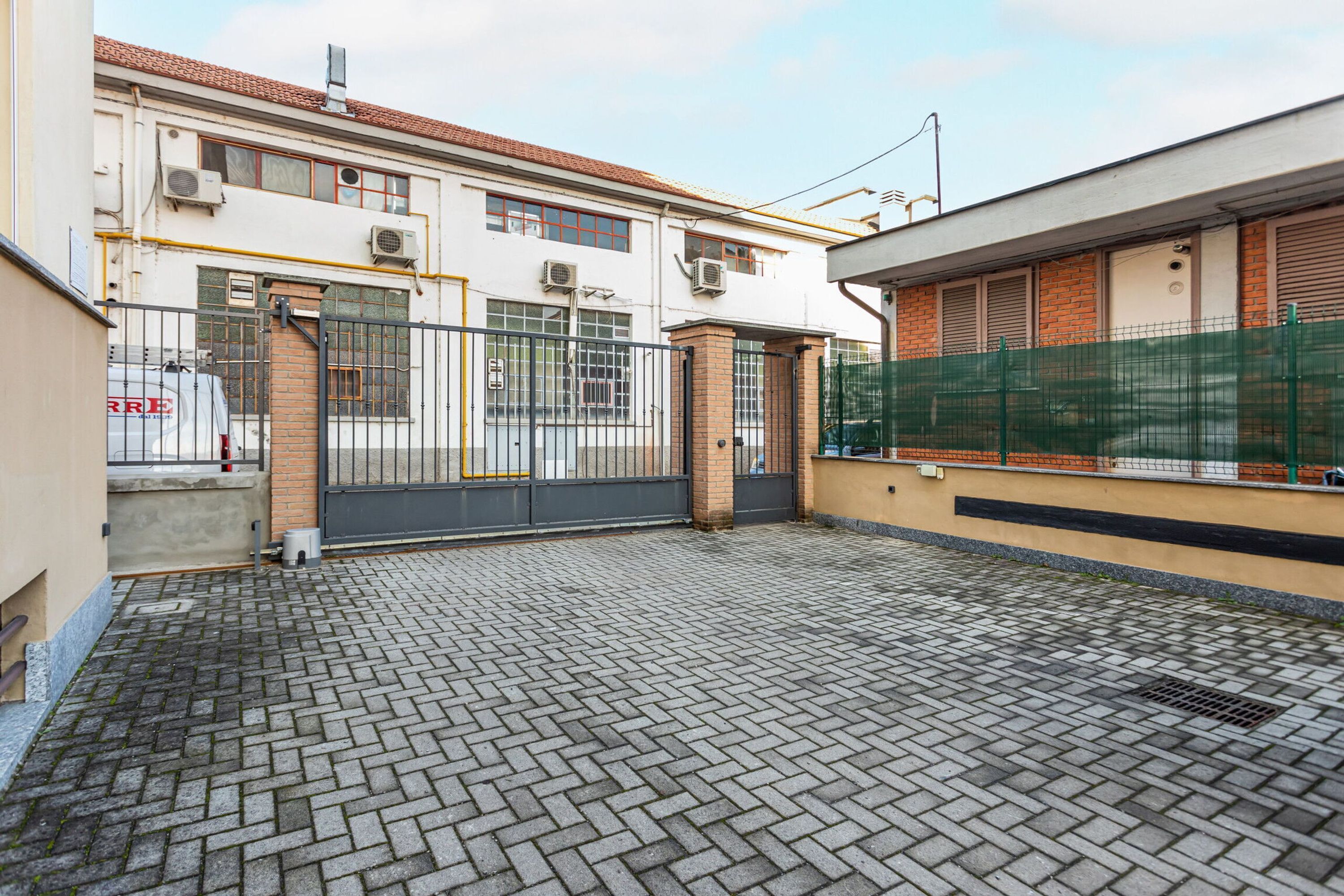 Une cour avec un sol pavé, une entrée fermée par un portail et un bâtiment doté de nombreuses fenêtres et de plusieurs unités de climatisation.