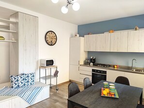 Eine moderne Küche mit weißen Schränken, einer Mikrowelle, einem Herd und einem Spülbecken. Ein Essbereich mit Tisch und Stühlen lädt zum Verweilen ein, und an der Wand hängt eine Uhr.