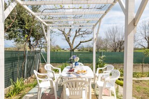 Eine überdachte Terrasse mit Tisch für zwei Personen, umgeben von weißen Plastikstühlen, einer Weinflasche und einer Obstschale.