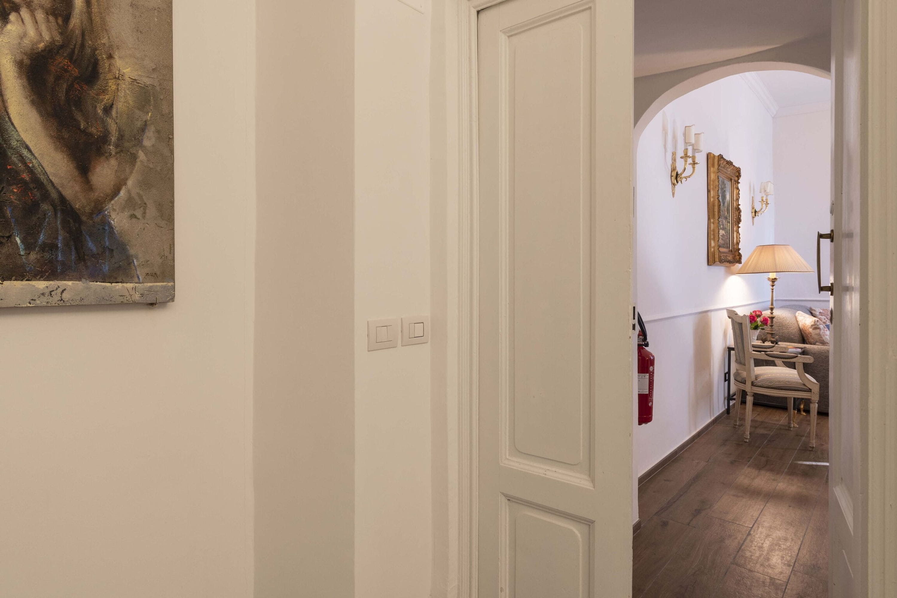 Un pasillo con una puerta blanca, un cuadro en la pared, una lámpara, una silla y un extintor.