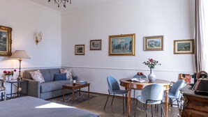 Quarto bem iluminado com sofá, mesa de jantar com cadeiras e cama. Há quadros nas paredes e um abajur em uma mesinha lateral.