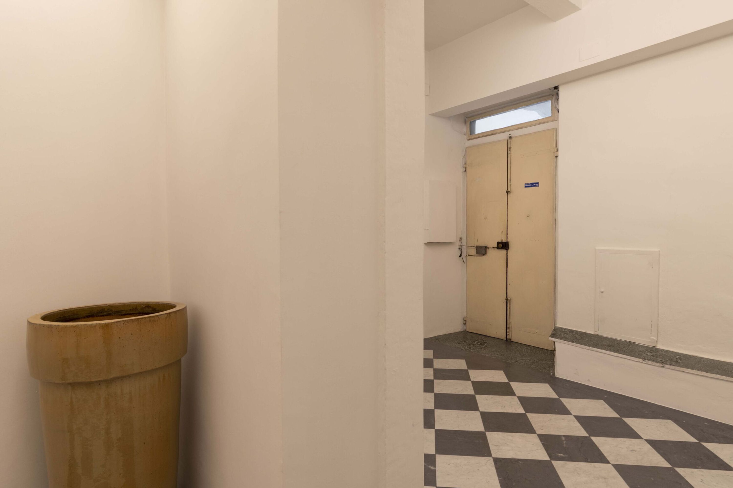 Un couloir étroit avec un sol à carreaux, une poubelle et une porte sur laquelle est apposée une pancarte.