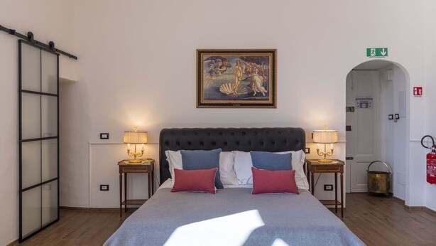 Quarto de hotel com uma cama grande, duas mesas de cabeceira com luminárias, um quadro acima da cama, uma porta deslizante e um sinal de saída de emergência.