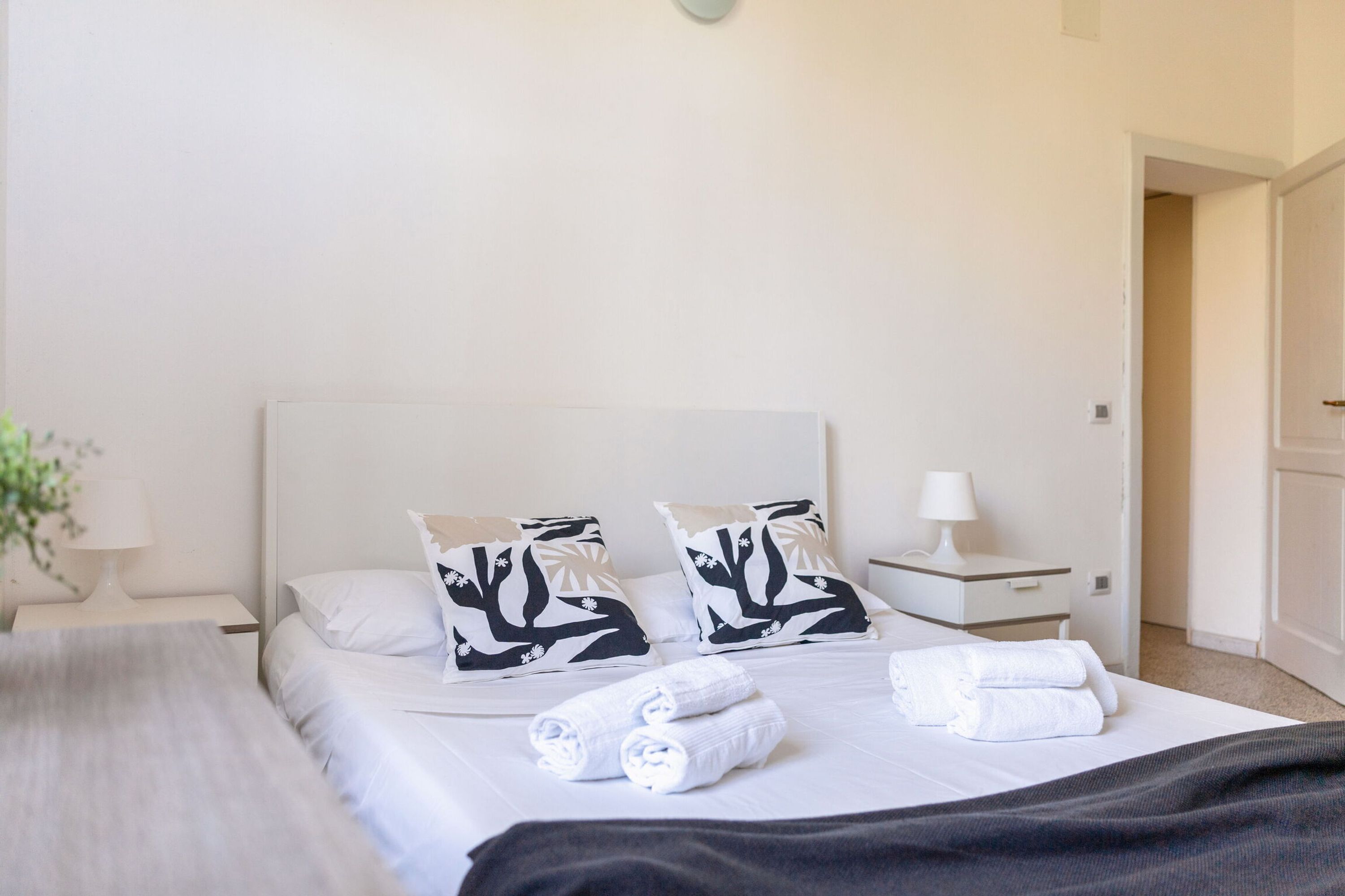 A hotel room with a bed, two pillows with a black and white pattern, a bedside table with a lamp, and a rolled towel on the bed.