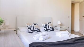 A hotel room with a bed, two pillows with a black and white pattern, a bedside table with a lamp, and a rolled towel on the bed.