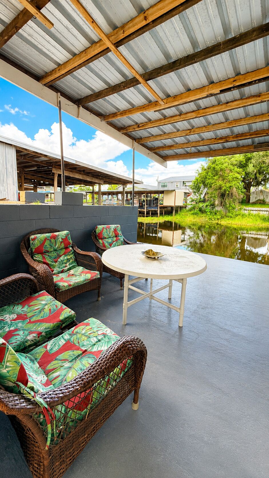 Un espace extérieur aménagé, abrité et doté de mobilier en osier, avec une table ronde, offrant une vue sur un plan d’eau et la verdure environnante.