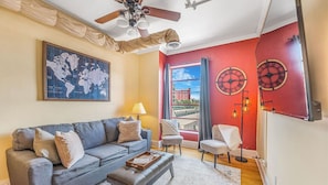 A living room with a sofa, a coffee table, a world map, a ceiling fan, and a red accent wall with clocks. - The Asheville Jewel Downtown Asheville 1BR with queen bed, full kitchen, Wi-Fi, and elevator. (Asheville)
