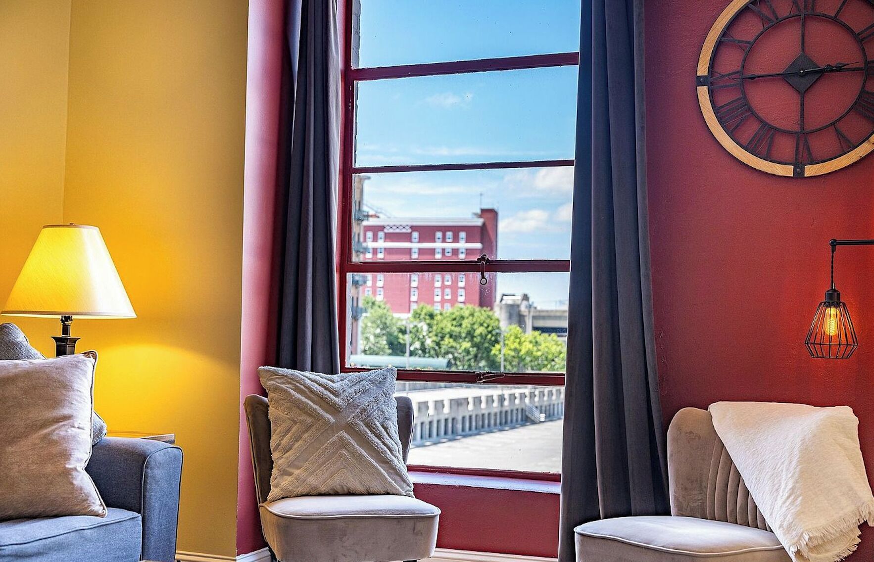 A room with a view of a red building, a clock on the wall, and a lamp on a table.