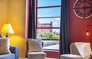 A room with a view of a red building, a clock on the wall, and a lamp on a table. - The Asheville Jewel Downtown Asheville 1BR with queen bed, full kitchen, Wi-Fi, and elevator. (Asheville)