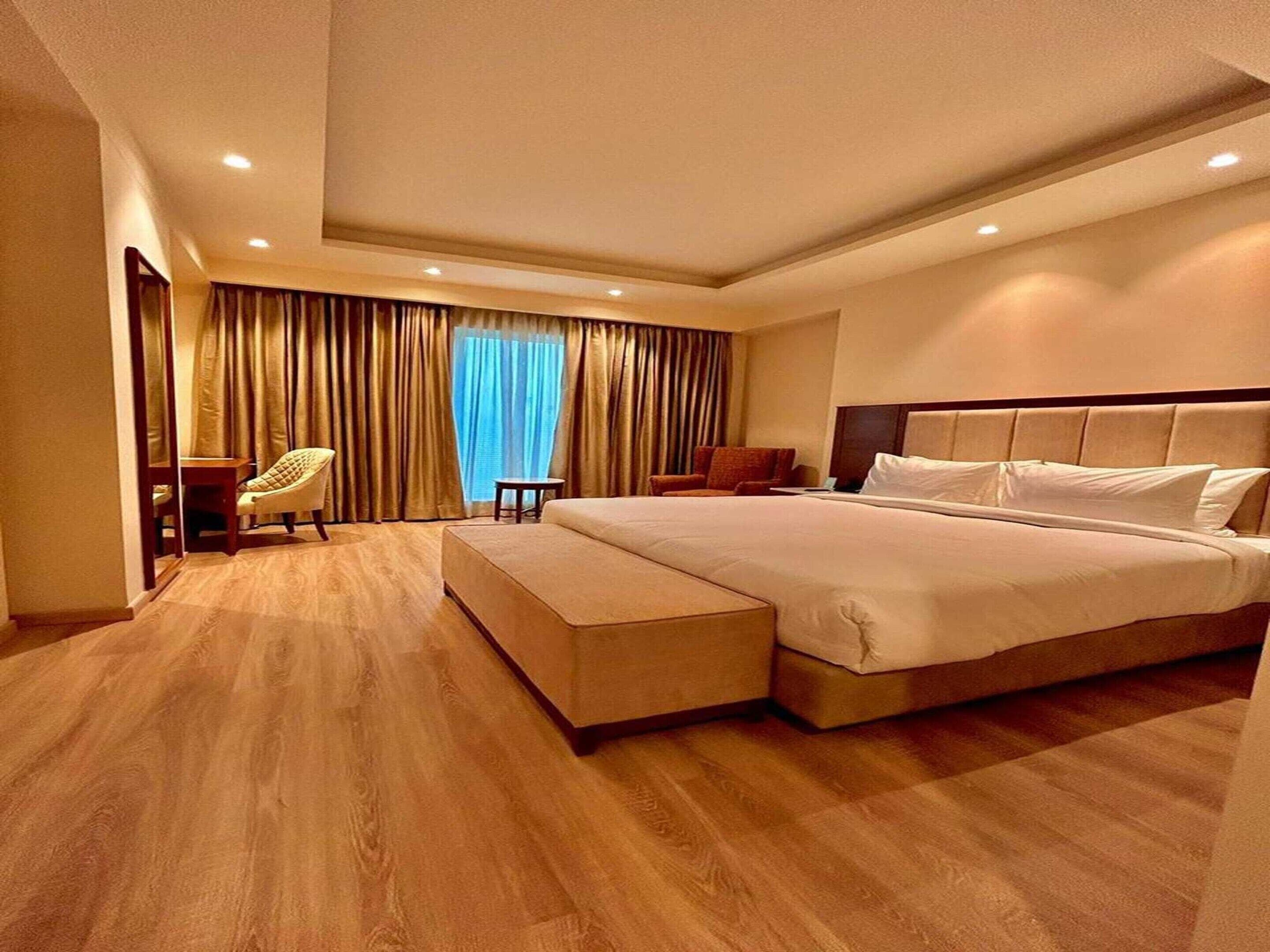 A hotel room with a large bed, a seating area with a chair, and a view of the outdoors through the window.