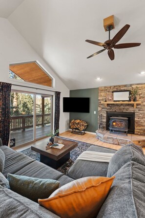 A spacious living room with a fireplace, a ceiling fan, and a large window overlooking a deck. - Private Mountain Retreat w/Hot Tub & Game Room I Sleeps 12 (Woodland Park)
