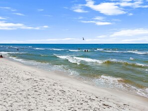 Una playa con olas, cielo despejado y un velero a lo lejos.