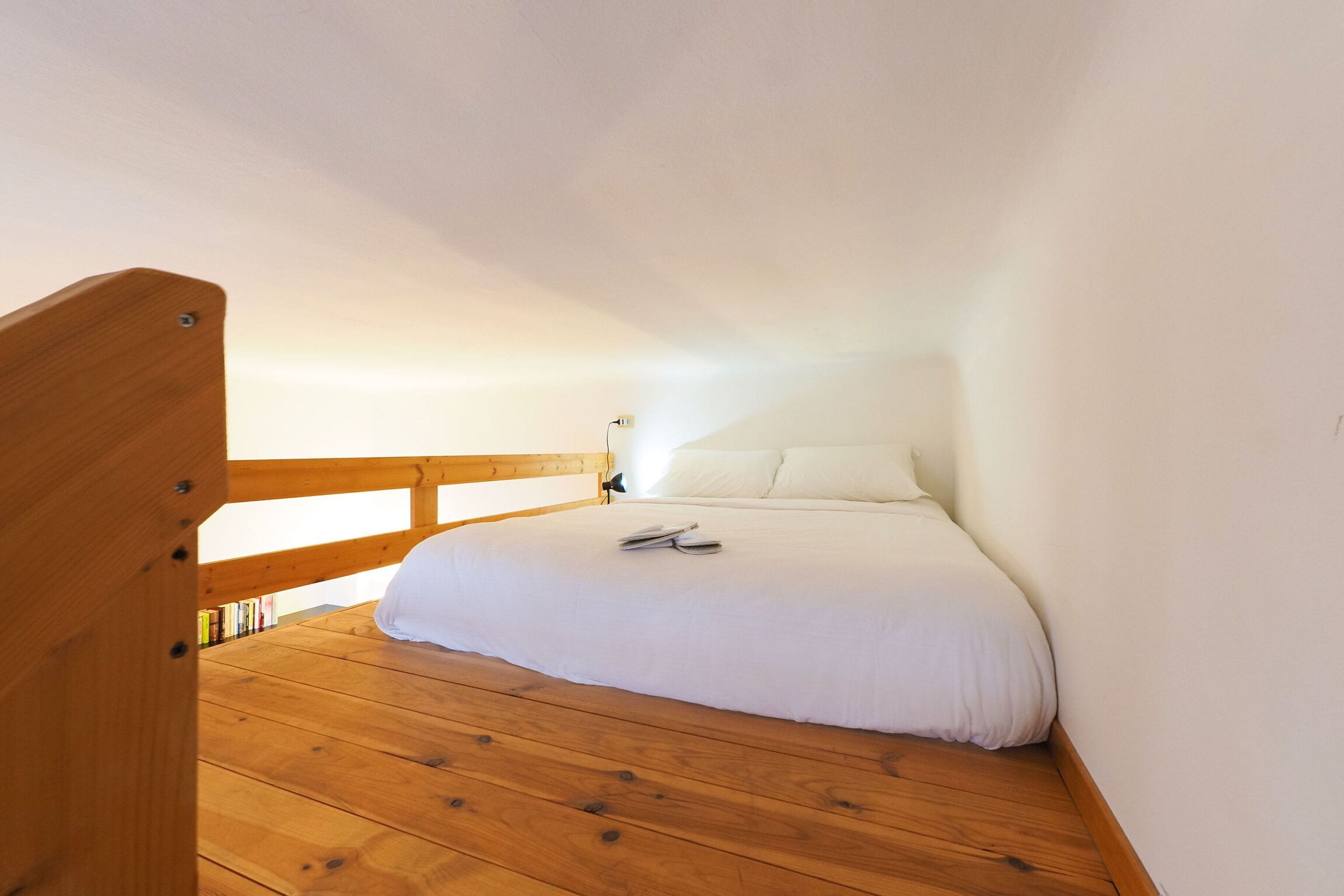 Un dormitorio con piso de madera, una cama con ropa blanca y una mesita de noche con lámpara.