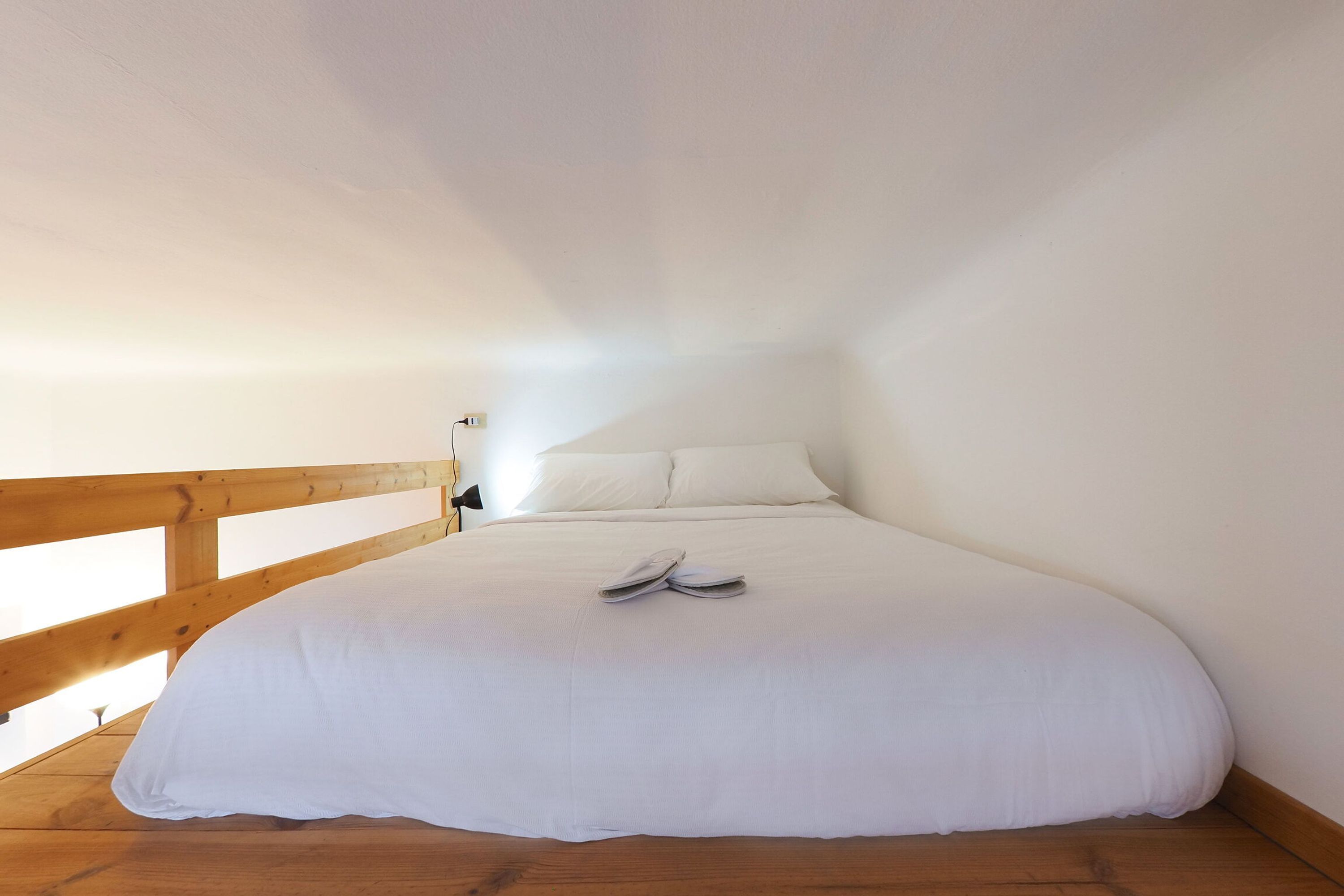 Habitació amb un llit individual, amb capçalera de fusta, roba de llit blanca i un parell de sabates sobre el llit.