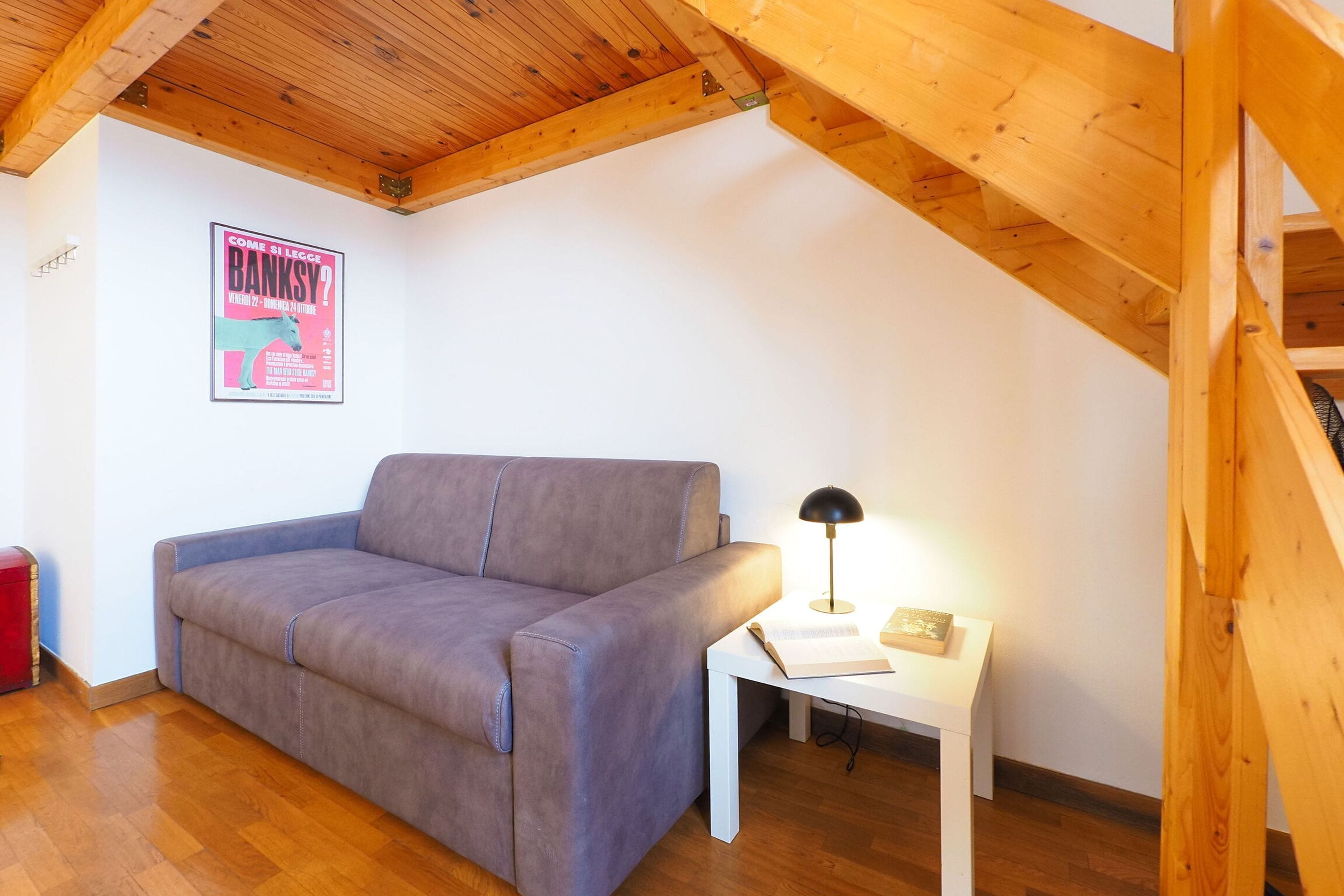 Una habitación con techo de madera, un sofá gris, una mesita de noche blanca con una lámpara y un cartel en la pared.