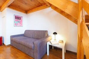 A room with a wooden ceiling, a grey sofa, a white side table with a lamp, and a poster on the wall. - Suite Meda in Milano (Milan)