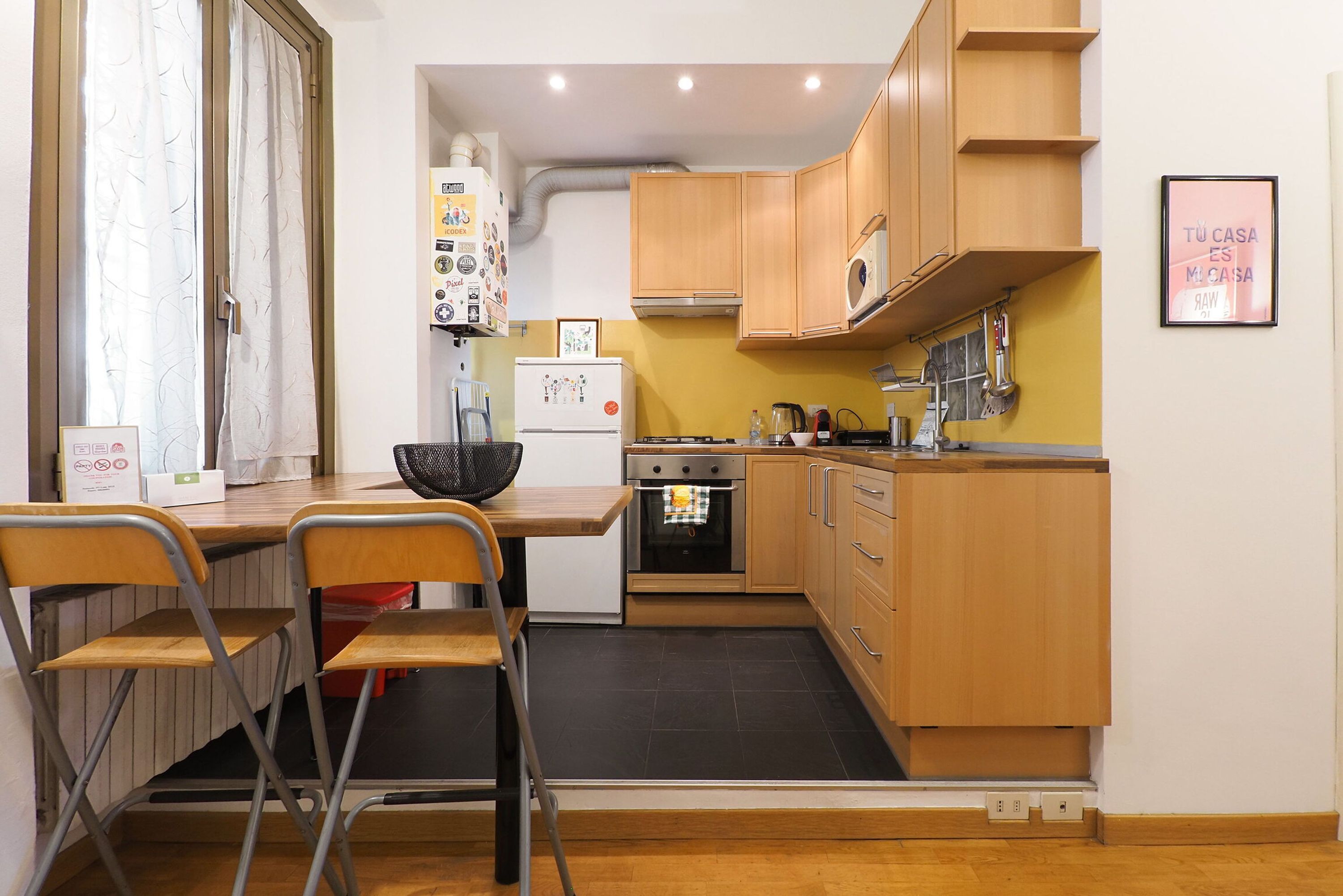 A compact kitchen with wooden cabinets, a built-in oven, and a microwave. There is a dining table with chairs and a window with curtains.