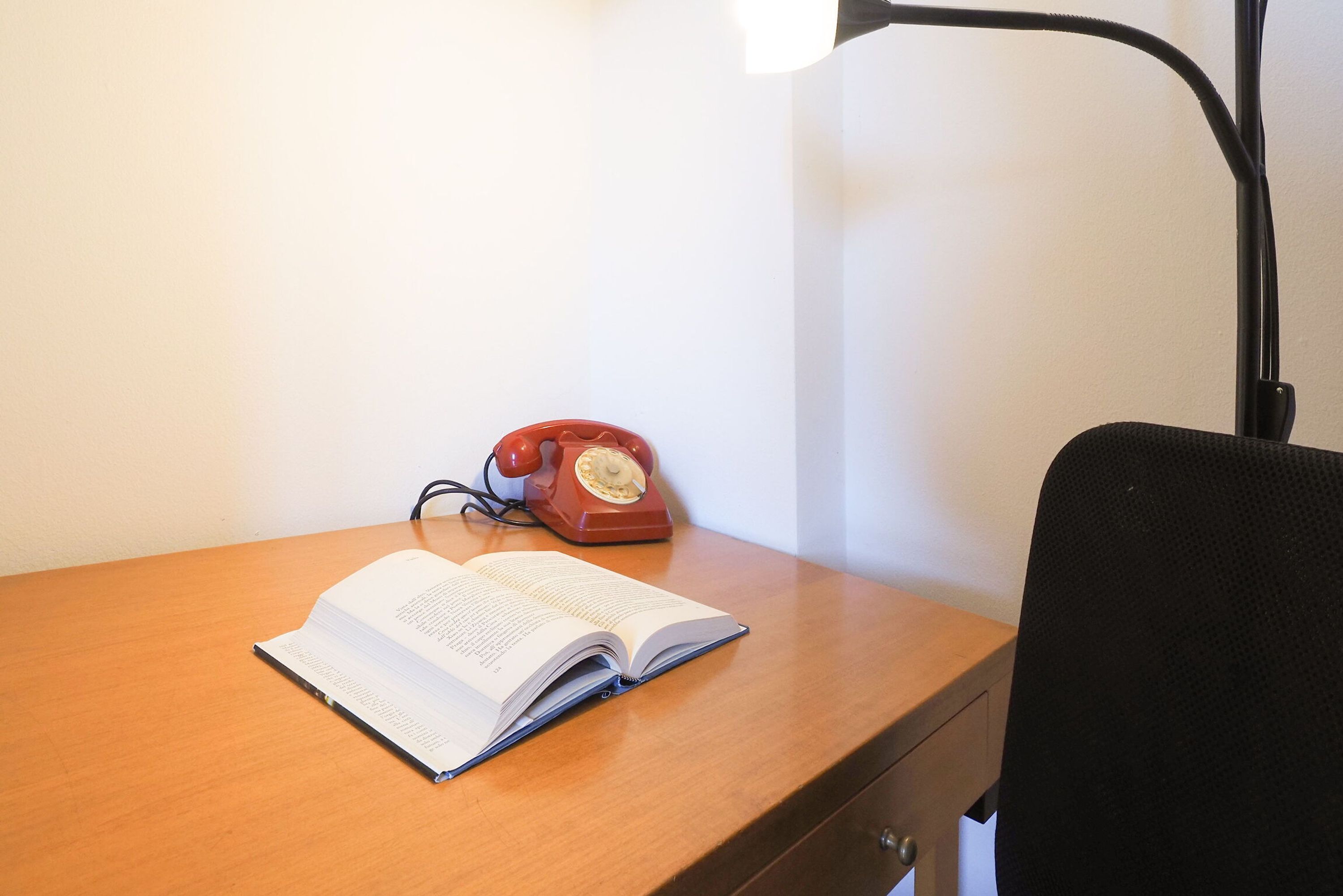 Een houten bureau met een open boek, een rode draaischijf telefoon en een zwarte bureaustoel.