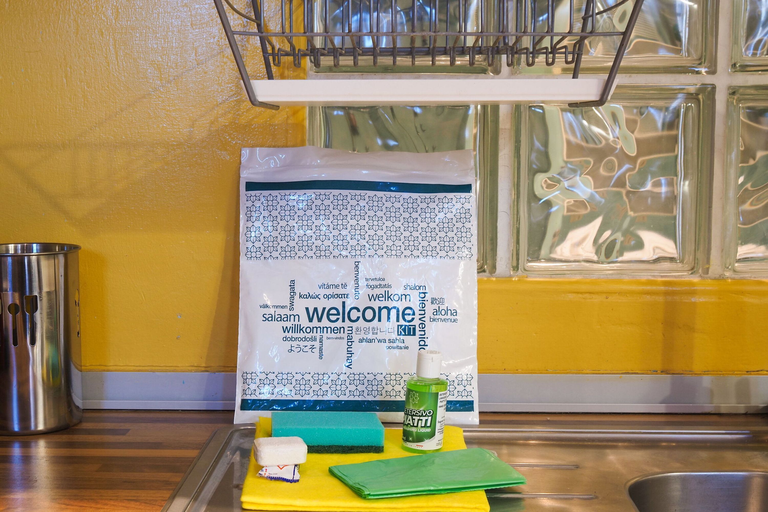 A kitchen with a yellow wall, a stainless steel sink, a metal rack, a bag of detergent, a green sponge, a white bottle, and a green cleaning liquid.
