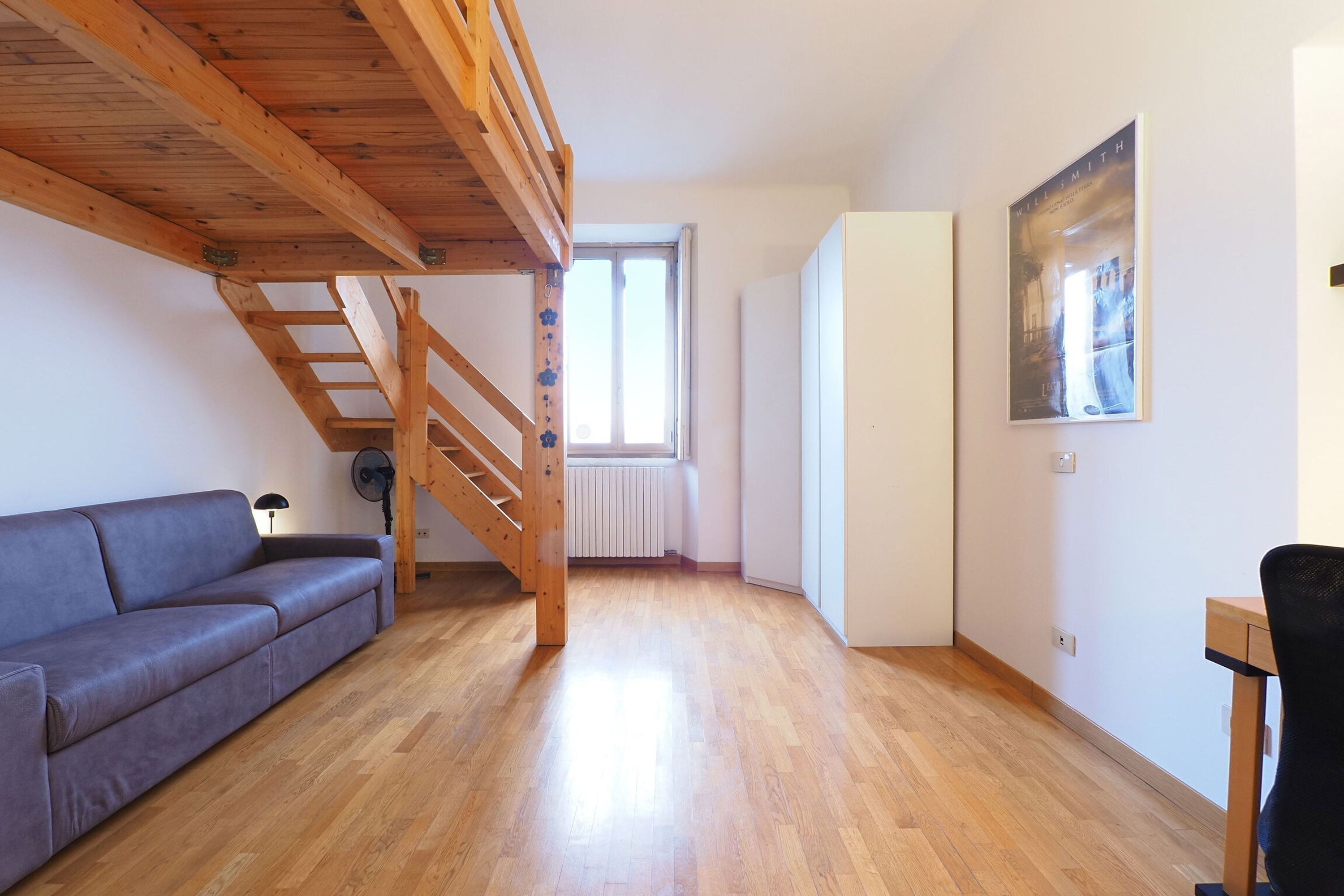 Una habitación con una escalera de madera, un sofá gris, un radiador y un cuadro enmarcado en la pared.
