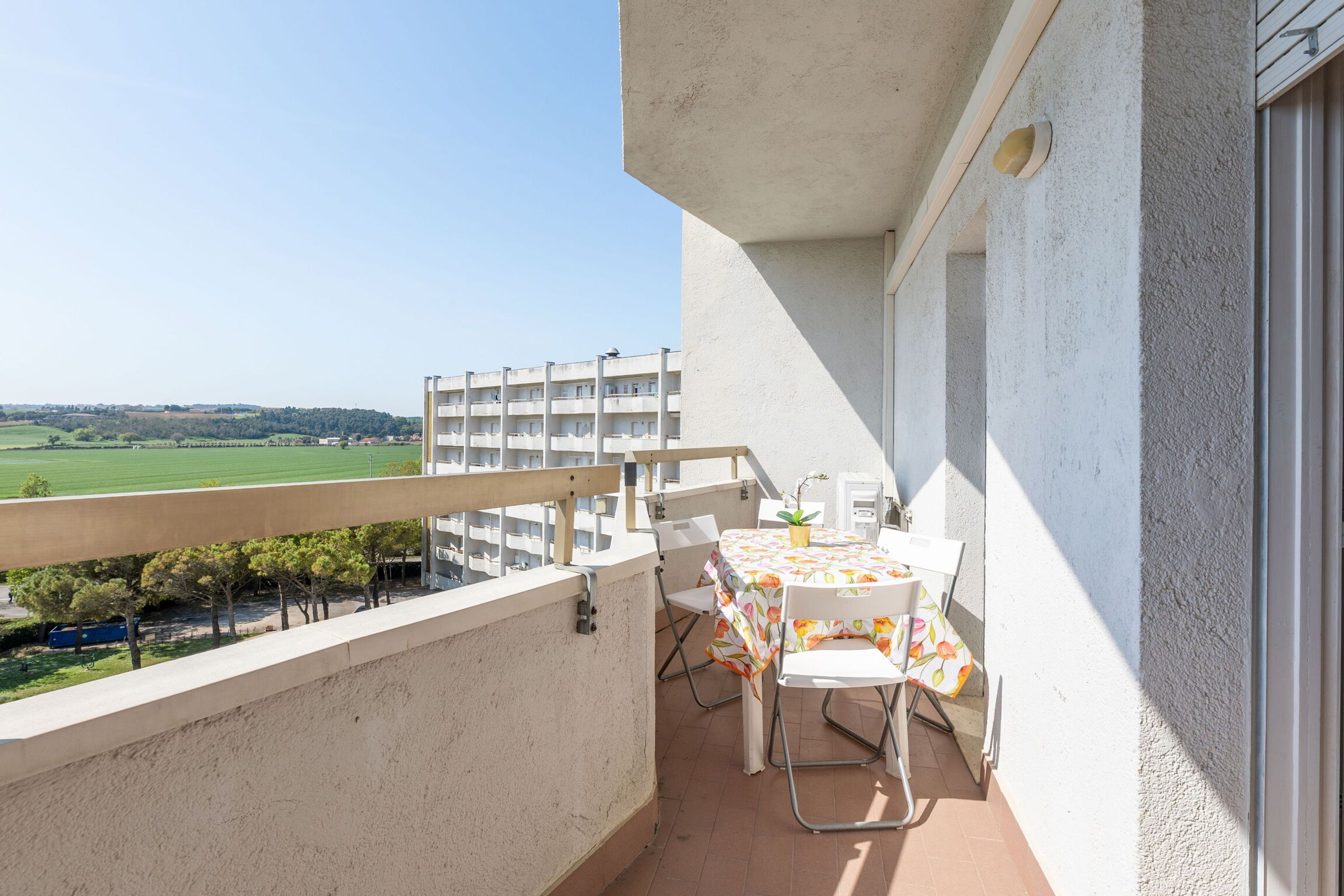 A balcony with a table and chairs, overlooking a green landscape and a building in the distance.