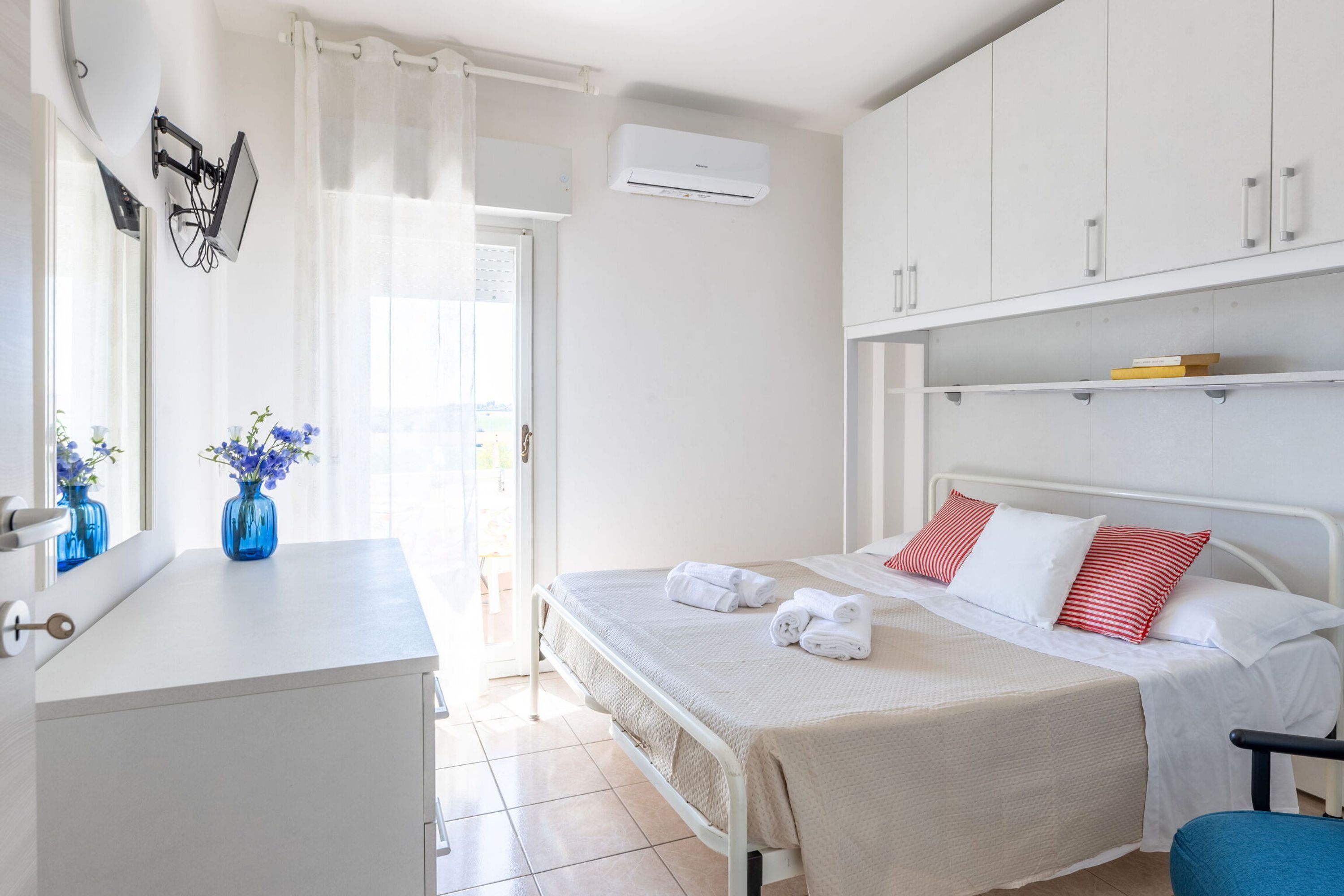 A bedroom with a bed, a bedside table with a vase, a window with curtains, a wardrobe, and an air conditioning unit.