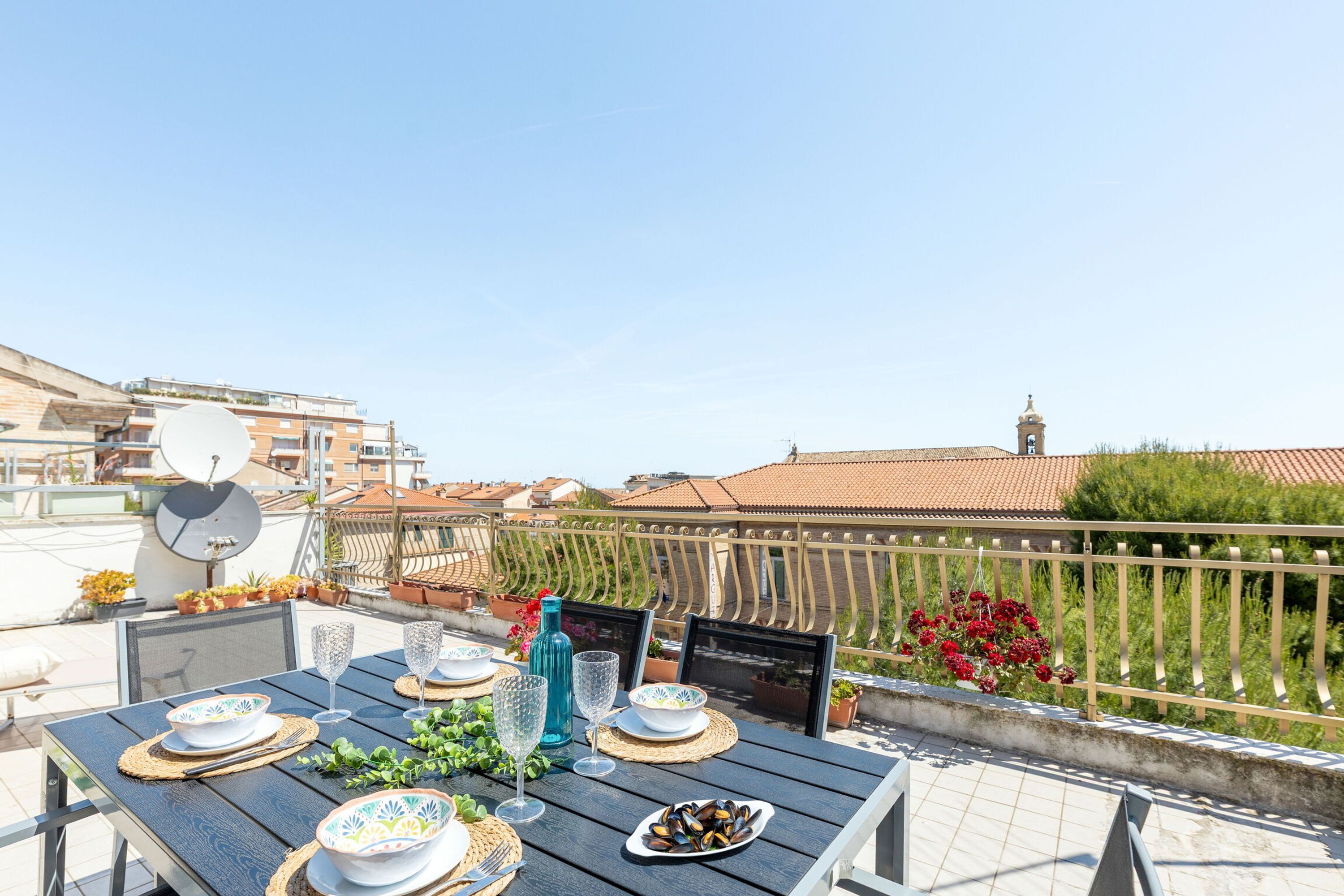 A rooftop terrace with a table set for dining, overlooking a cityscape with buildings and a bell tower.