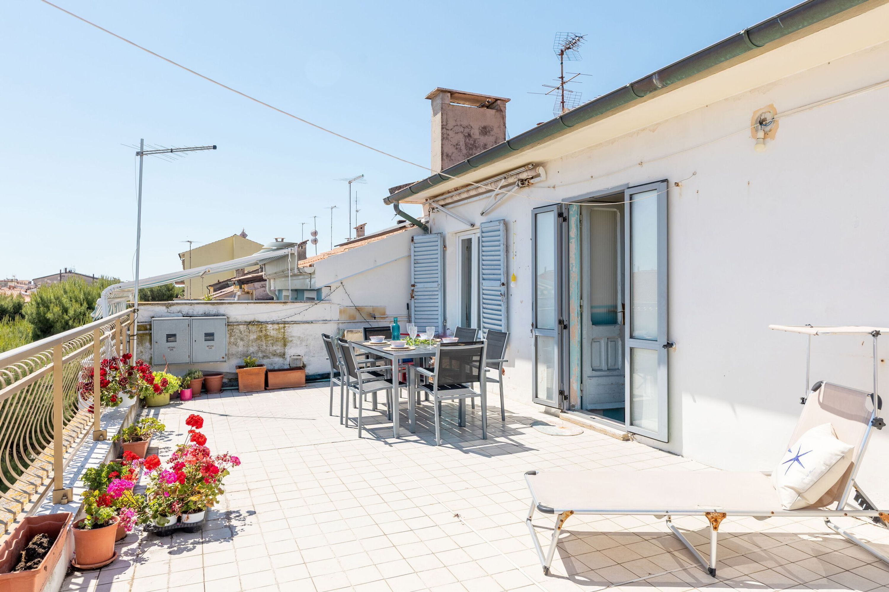 A rooftop terrace with a dining table, chairs, potted plants, and a sun lounger.