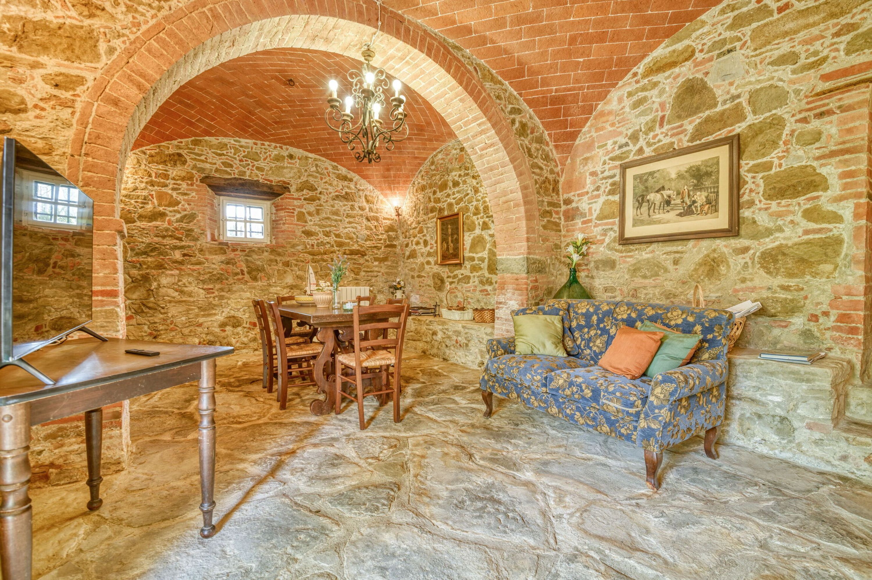 A stone-walled room with a dining table, chairs, a sofa, and a chandelier.