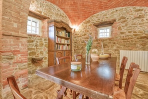 Una habitación rústica con paredes de piedra, una mesa de madera, sillas, una estantería y una ventana.