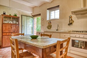 Una cocina con encimera de mármol, sillas de madera, un tazón verde, un armario de madera y un ventanal con vistas a la vegetación.
