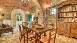 Un interior rústico con una pared de piedra, una mesa de comedor de madera, sillas, una estantería y una araña.