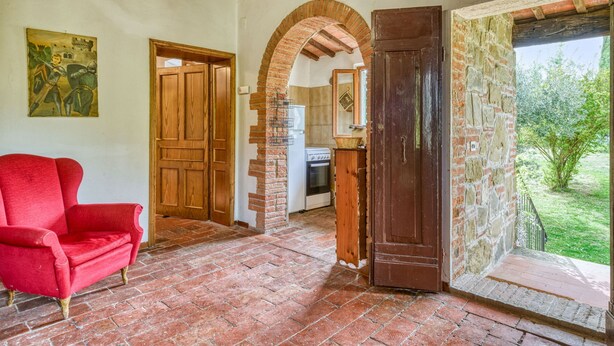 Una habitación con un sillón rojo, puertas de madera, un arco de ladrillo, una zona de cocina y vistas al exterior con vegetación.