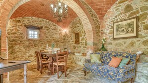 Un interior rústico con paredes de piedra, una mesa de comedor de madera con sillas, un sofá estampado y una araña.