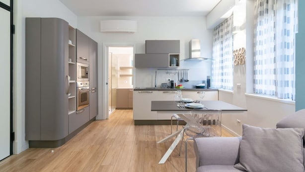 A modern kitchen with grey cabinets, a built-in oven, and a dining area with a glass table and chairs.
